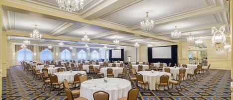A large banquet hall with round tables and chairs, chandeliers, and a carpeted floor.