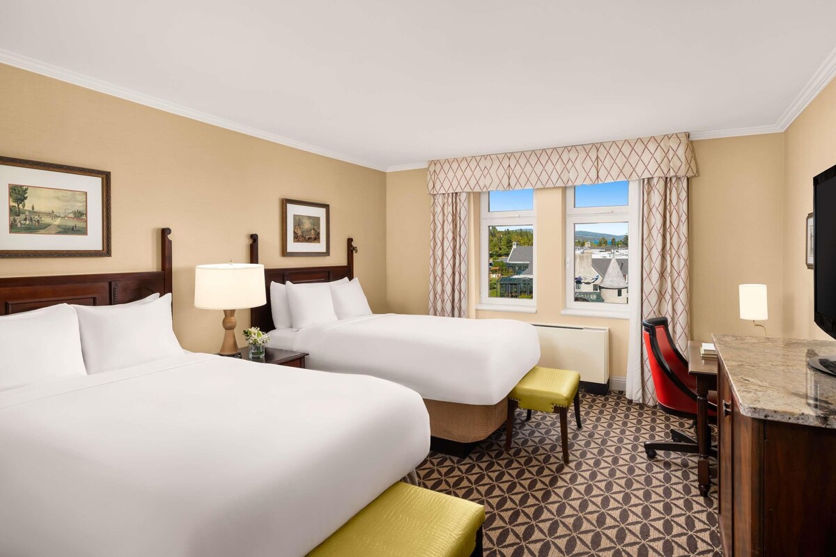 A hotel room with two beds, a desk, a chair, a TV, and a window with a view.