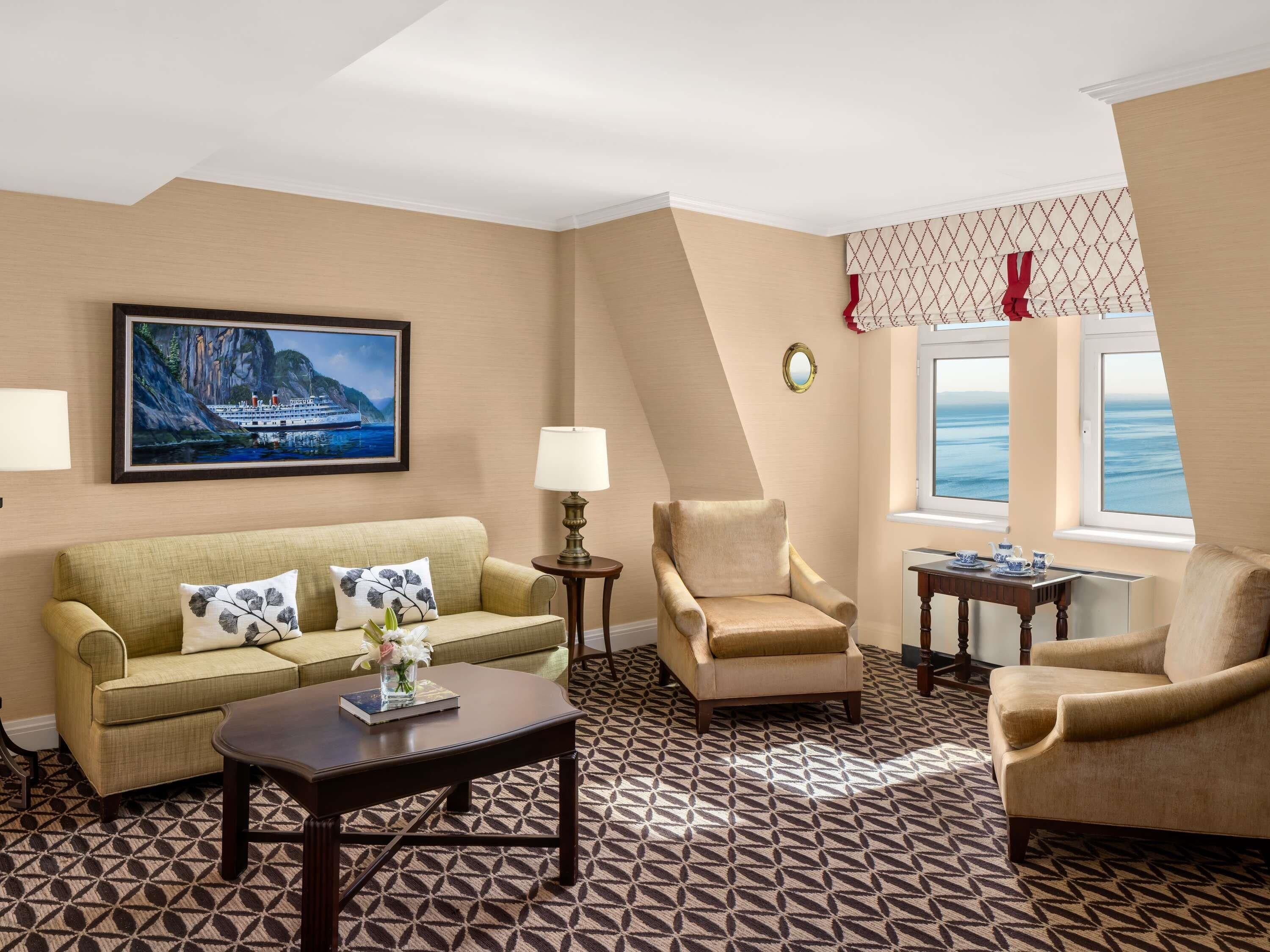 A hotel room with a sofa, armchairs, a coffee table, a lamp, a painting, a window with a view, and patterned carpet.
