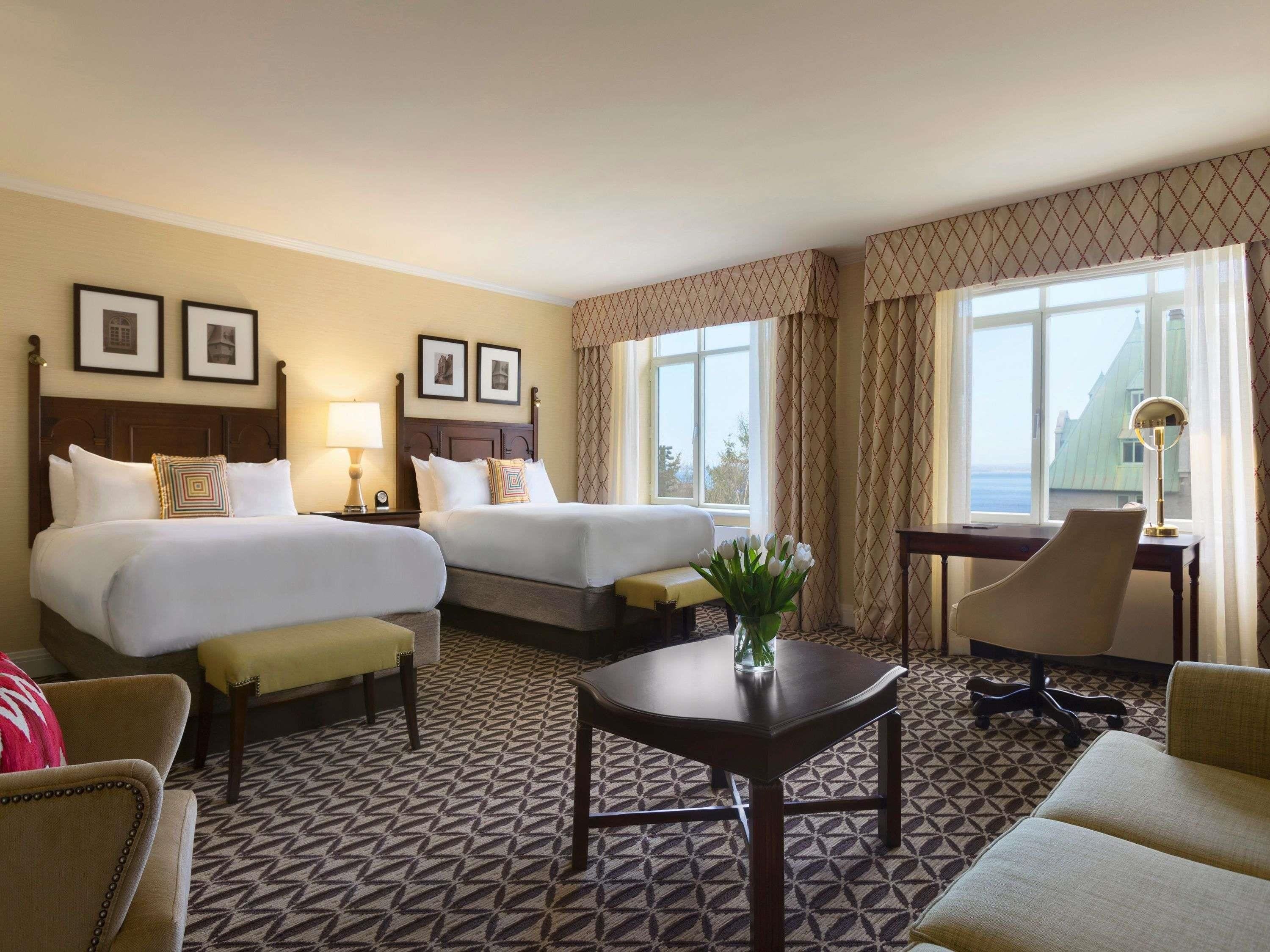 A hotel room with two beds, a desk, a chair, and a view of the sea.