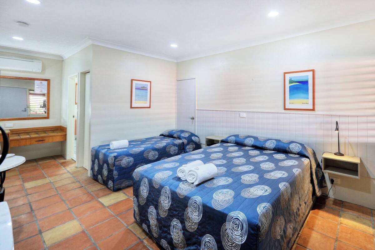 A hotel room with two beds, a desk, a chair, a mirror, a window with blinds, and a wall-mounted shelf.