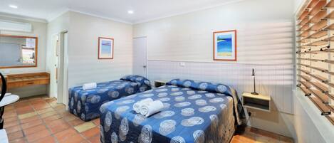 Une chambre d’hôtel avec deux lits, un bureau, une chaise, un miroir, une fenêtre avec des stores et une étagère fixée au mur.