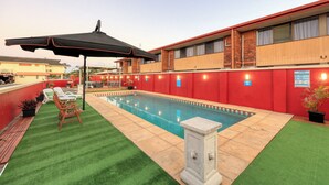 A swimming pool with a red wall and a covered seating area with chairs and a table.