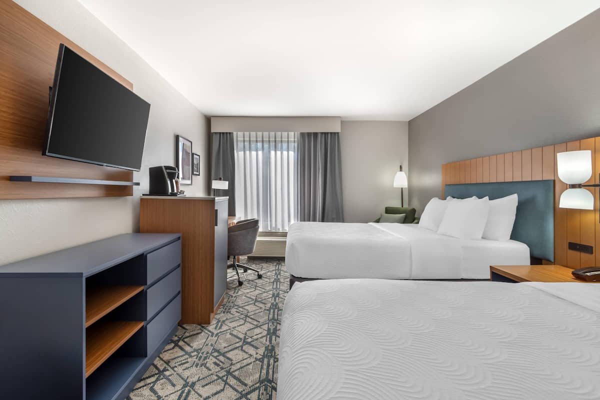 Une chambre d’hôtel équipée d’une télévision à écran plat, d’un bureau avec une chaise, de deux lits, d’une tête de lit avec une lampe et d’une fenêtre avec des rideaux.