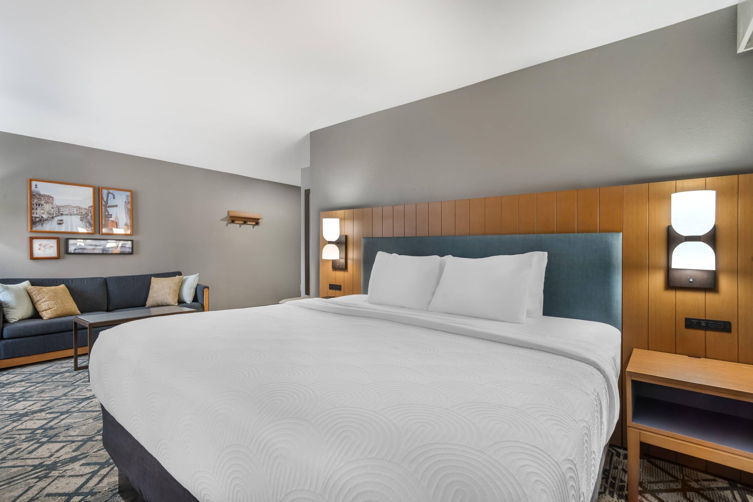 A hotel room with a large bed, a wooden headboard, two bedside tables, a sofa, and wall-mounted lights.