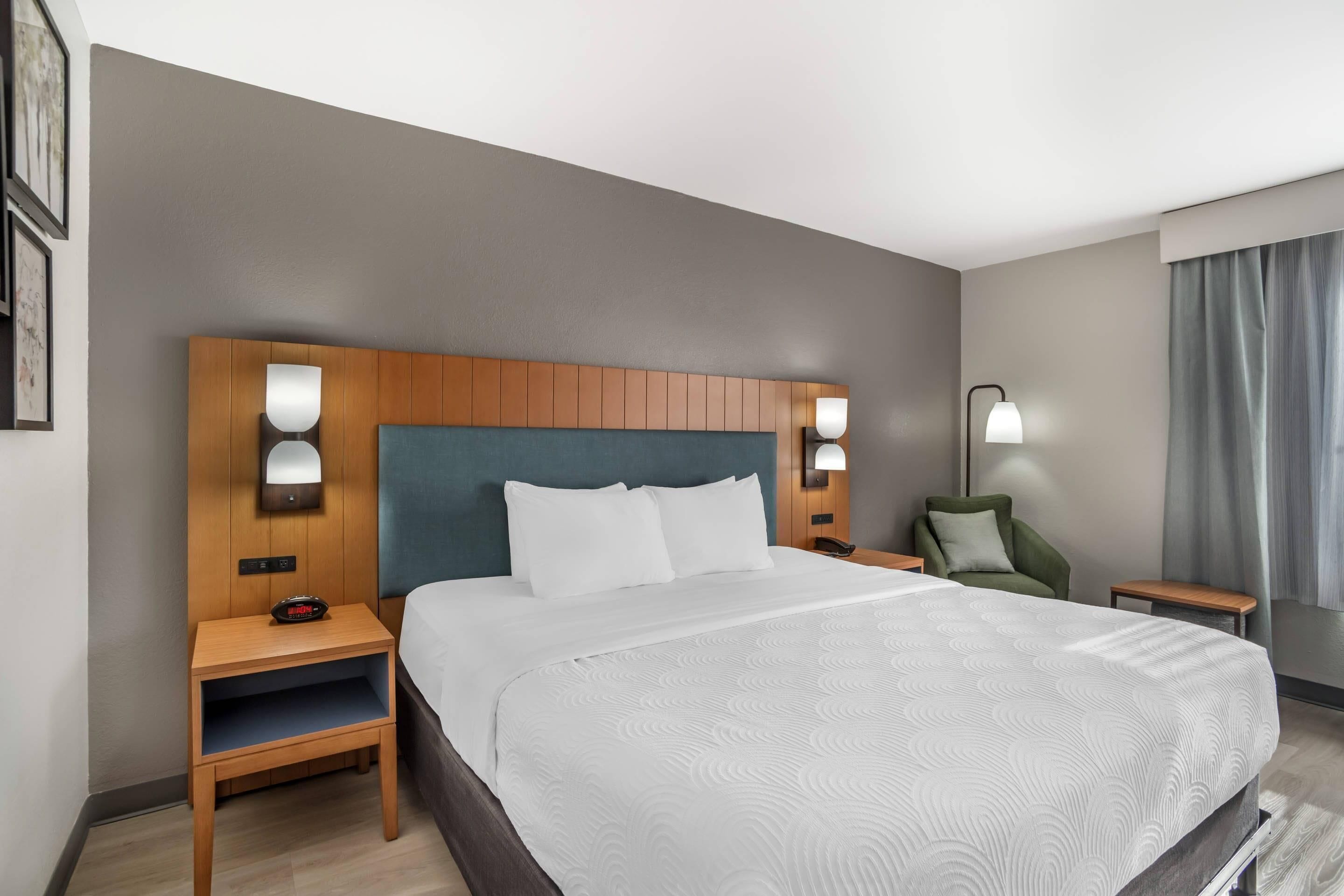 A hotel room with a large bed, wooden headboard, two bedside tables with lamps, a green chair, and a view of the outdoors.