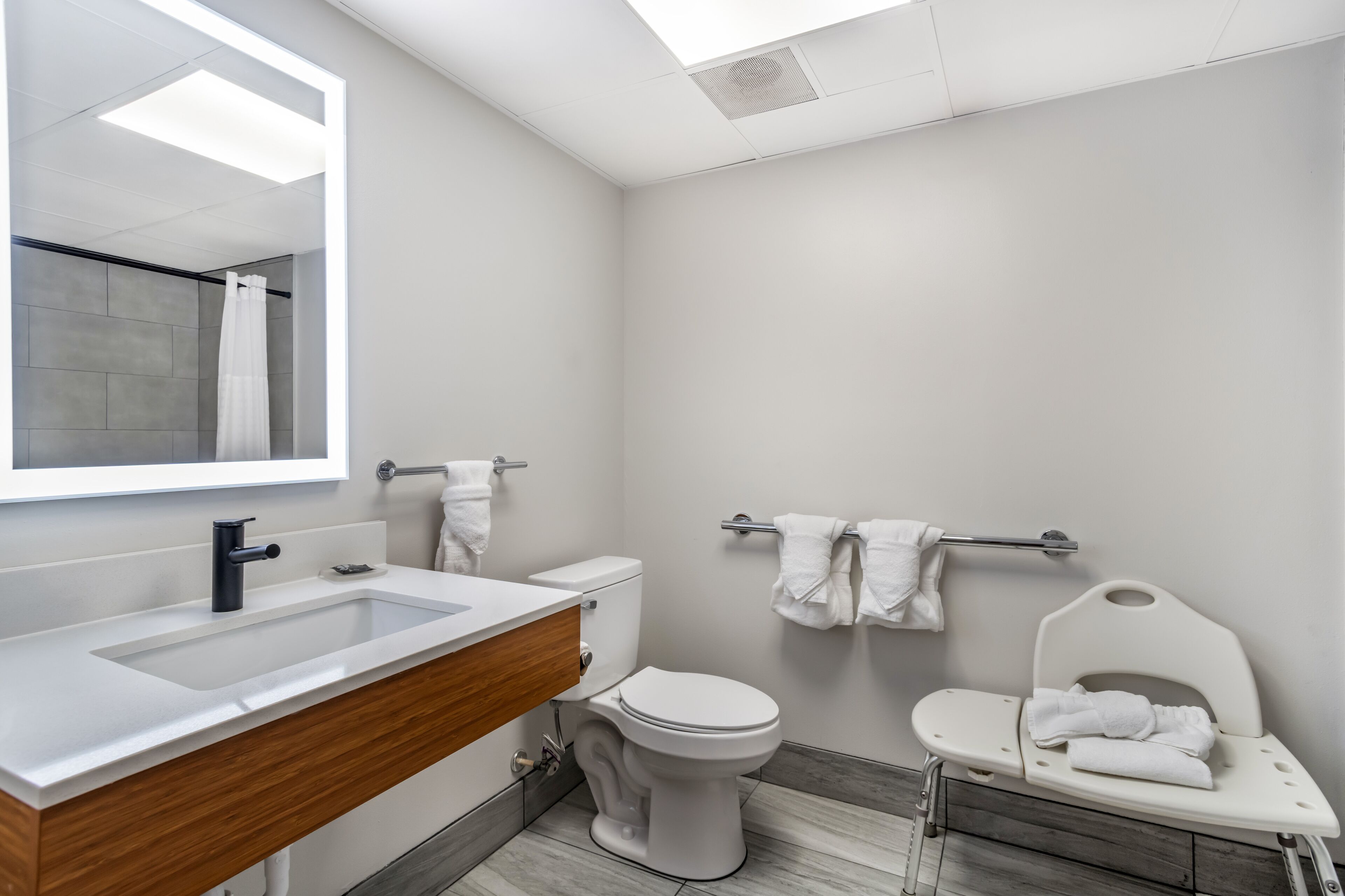 A modern bathroom with a large mirror, white sink, and toilet. There is a shower area with a curtain and a towel rack.