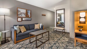 A modern hotel room with a grey sofa, a coffee table, a patterned carpet, a small side table, a bench, and a wall-mounted TV.