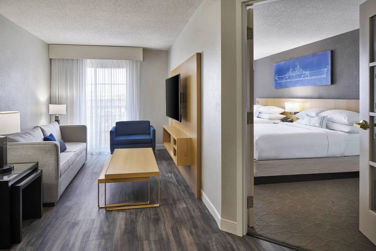 A hotel room with a sofa, a TV, a wooden coffee table, a bed with white bedding, and a wall-mounted TV.