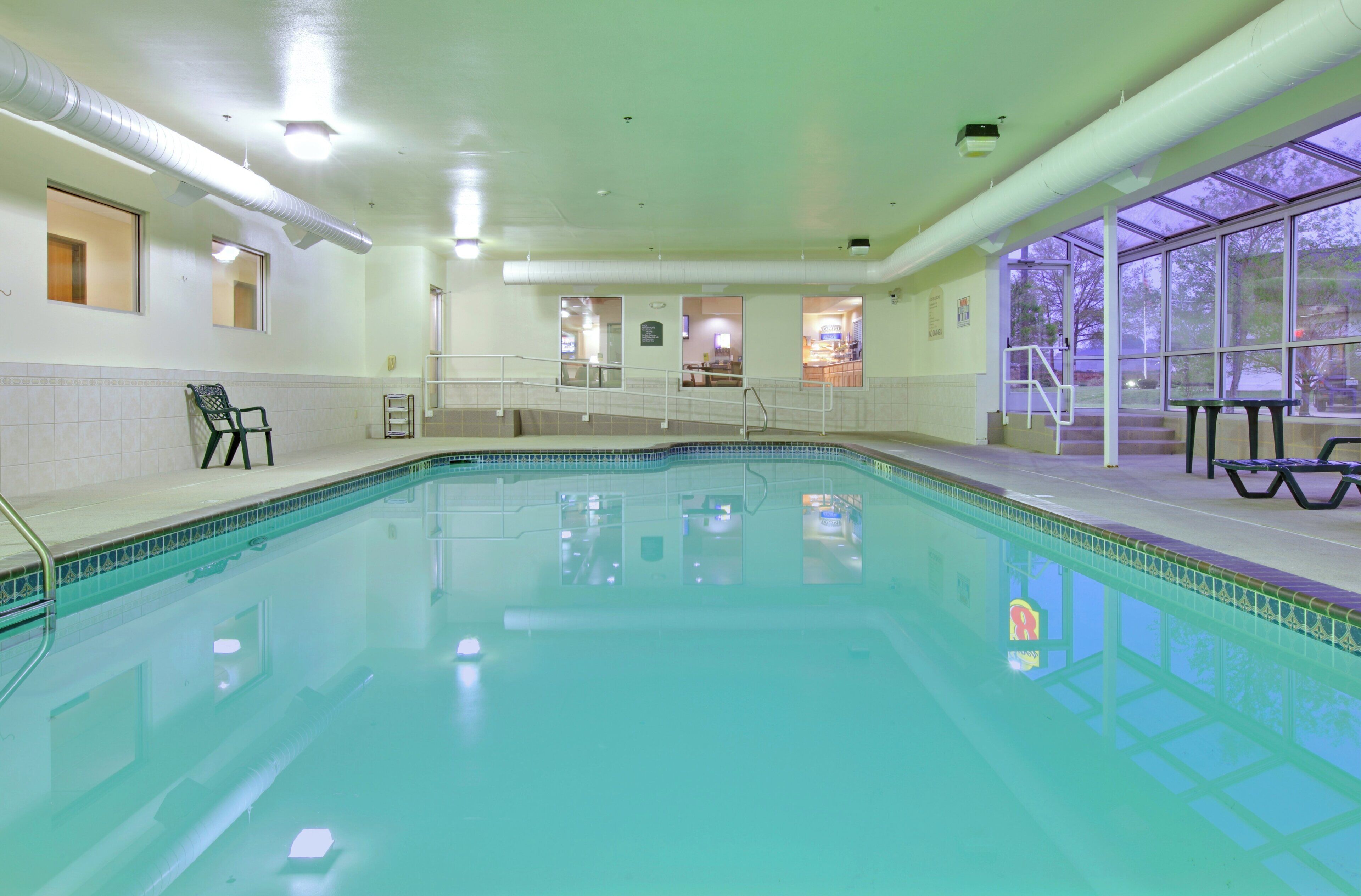 An indoor swimming pool with clear water, a tiled deck, and a view of the outside through large windows.