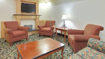 A hotel room with a flat-screen TV mounted above a fireplace, two armchairs, a sofa, a small table with a lamp, and a patterned carpet.