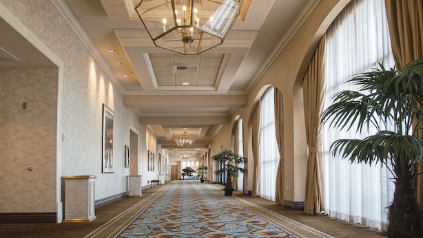 A long, narrow hallway with a patterned carpet, large windows with curtains, and a chandelier.
