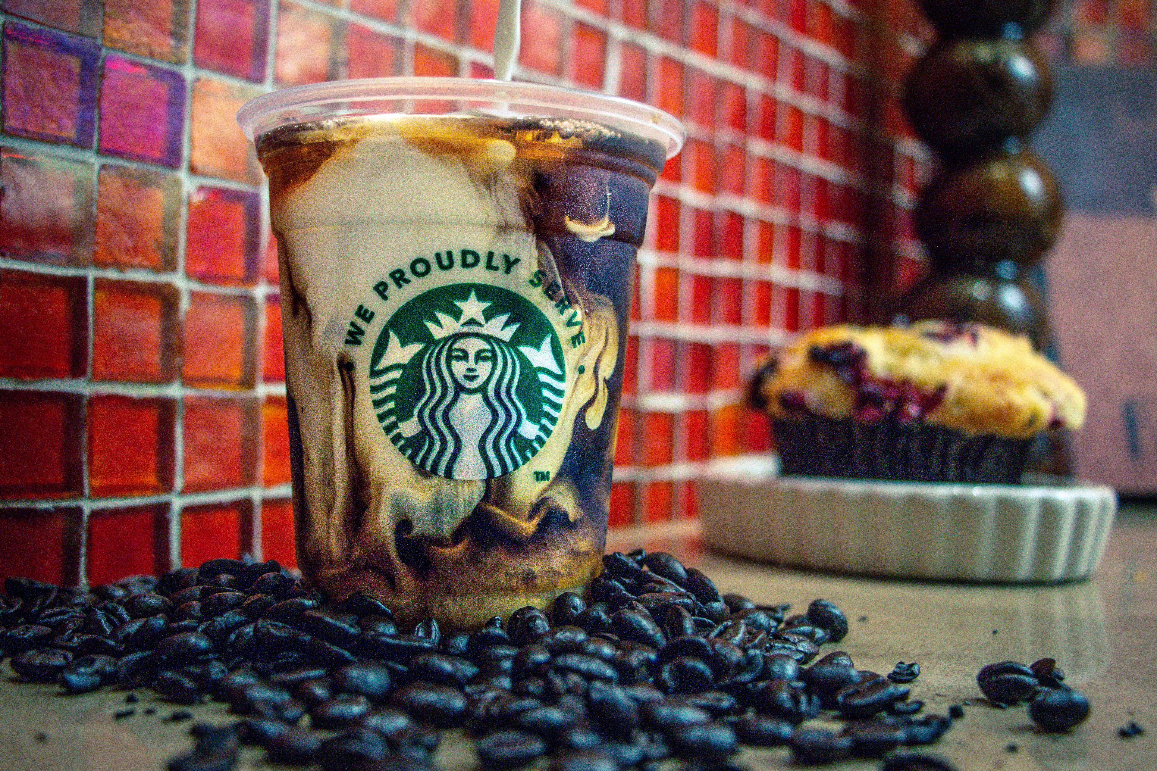 A Starbucks cup with iced coffee and a muffin on a table with coffee beans.