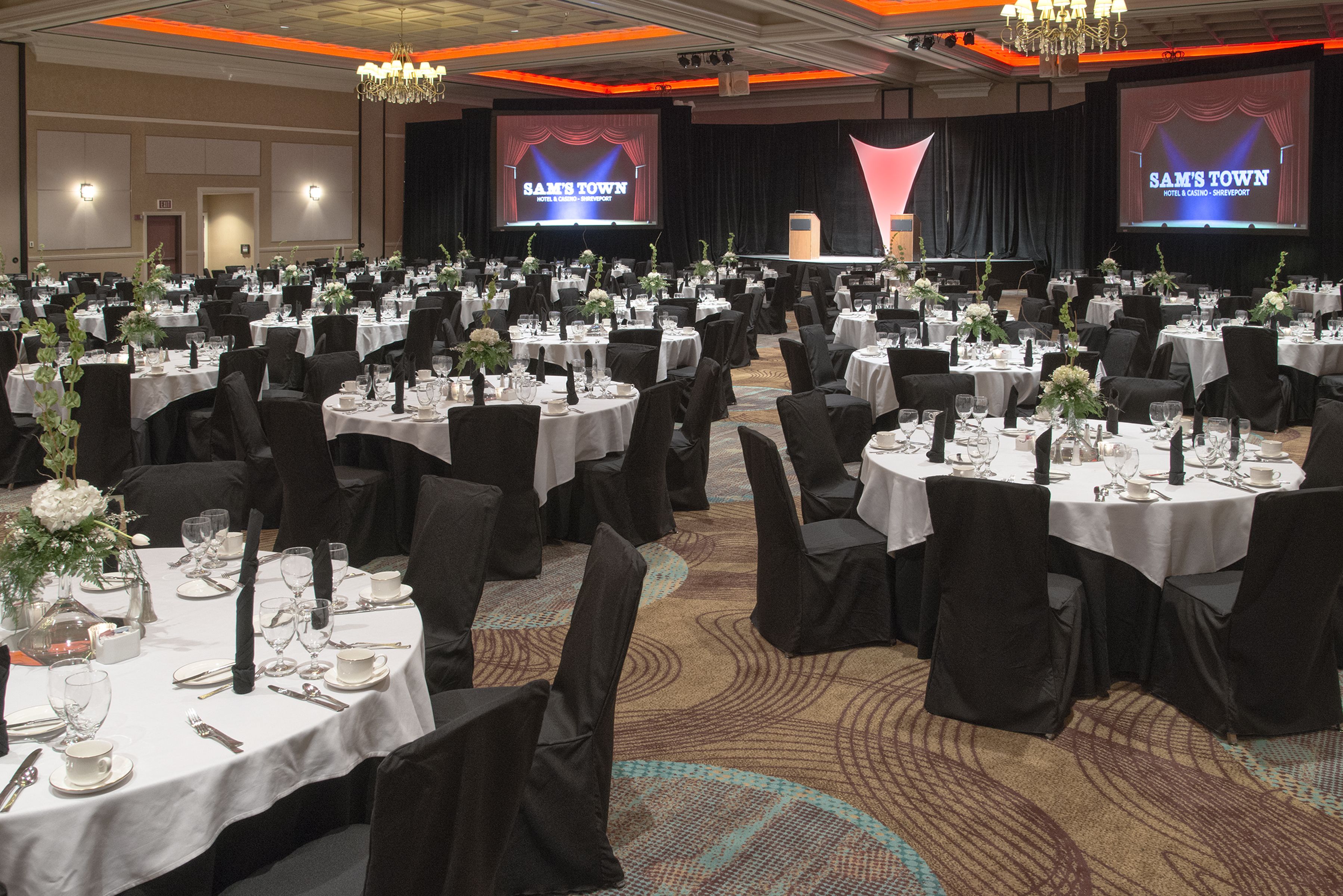 Un salón de banquetes con mesas redondas dispuestas para un evento formal, con manteles blancos, sillas negras y centros de mesa florales.