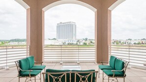 A balcony with two green cushioned chairs and a small table, overlooking a cityscape with a tall building.