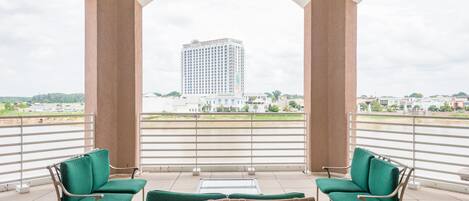 A balcony with two green cushioned chairs and a small table, overlooking a cityscape with a tall building.