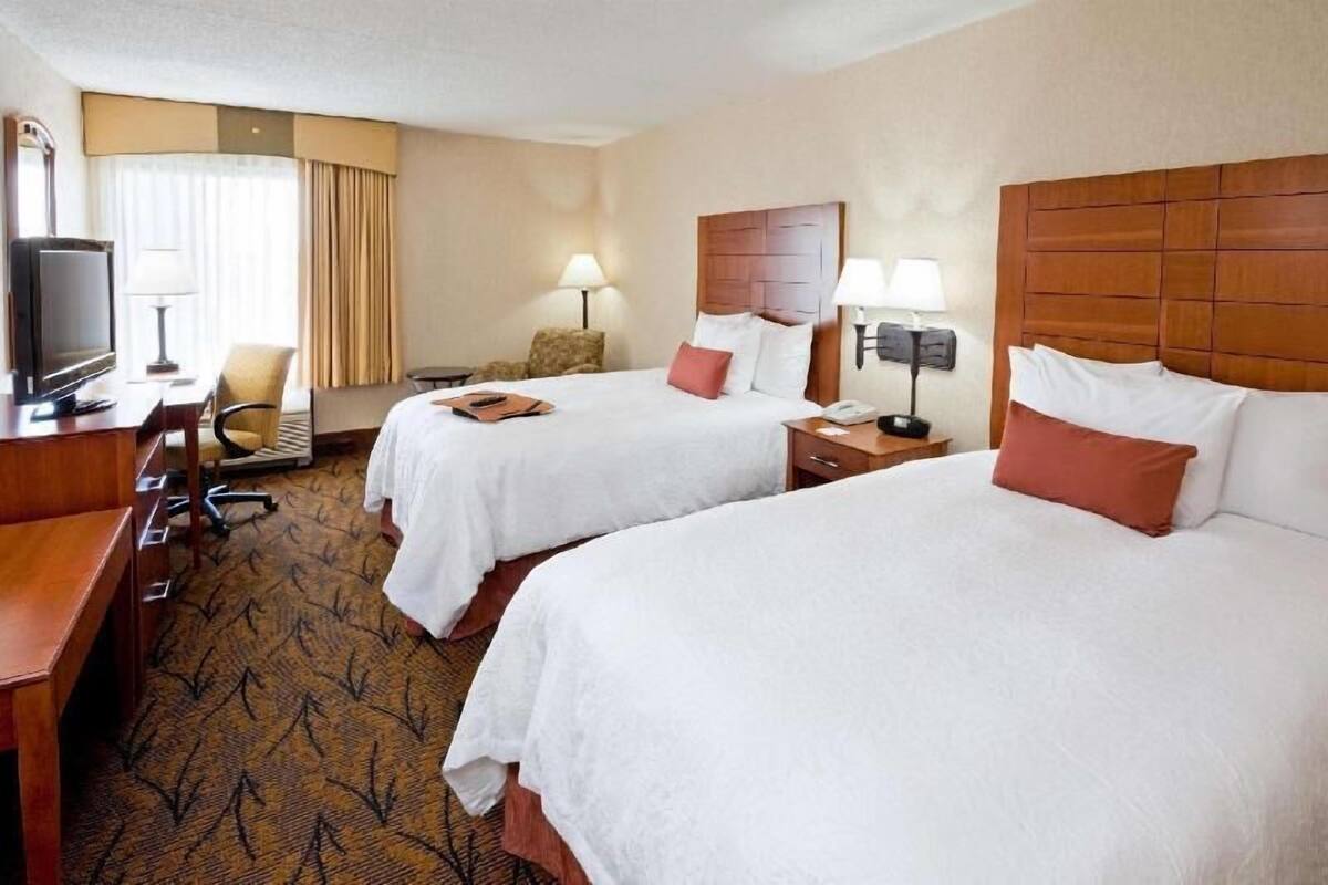 A hotel room with two beds, a desk with a television, lamps, and a carpeted floor.