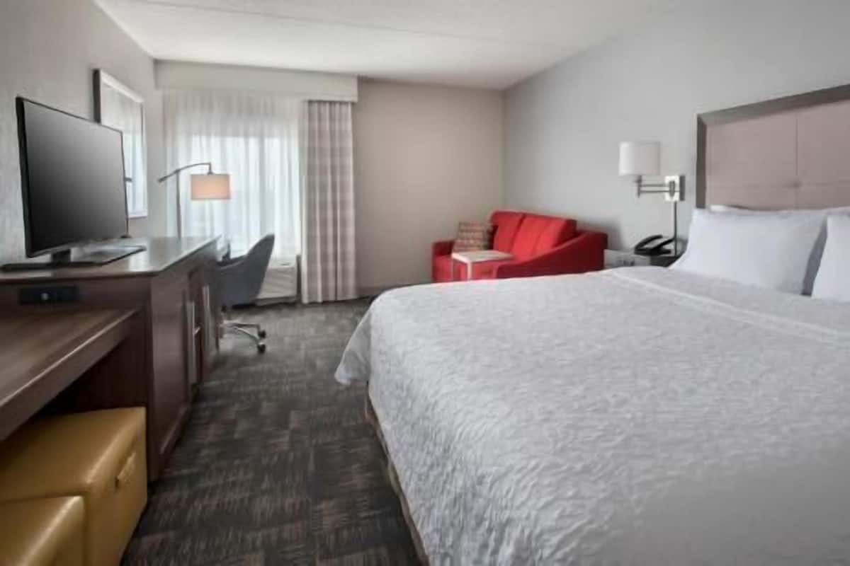 A hotel room with a bed, a desk with a computer, a TV, a red chair, and a lamp.