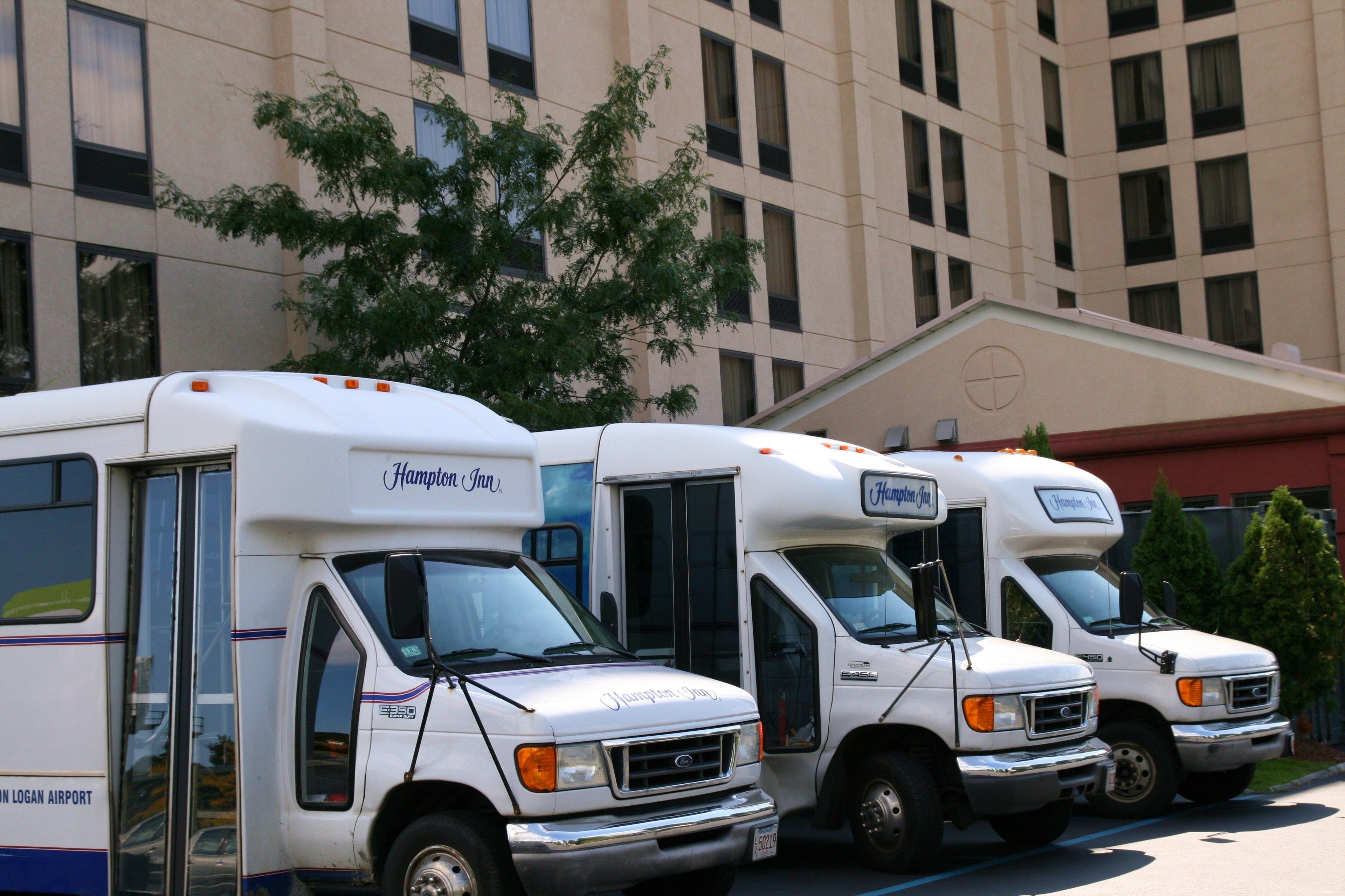 Three white motorhomes parked in a row in front of a building with a large tree and a red awning.