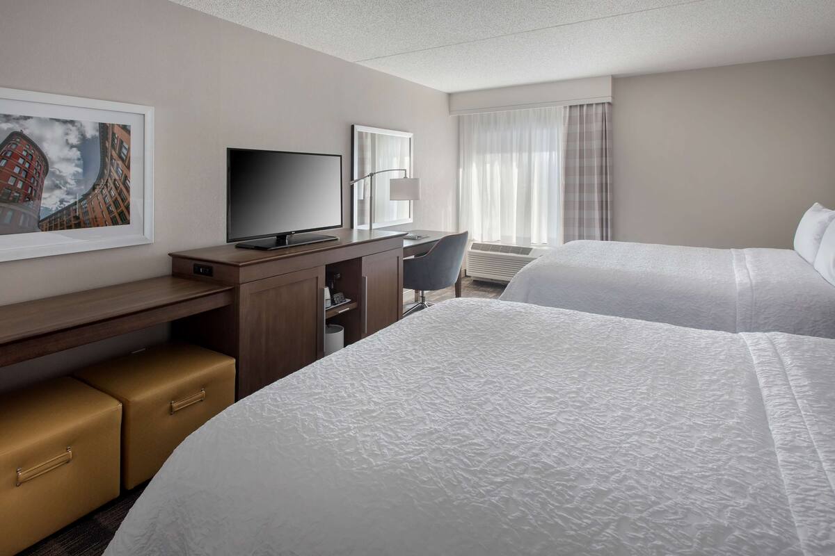 A hotel room with two beds, a desk with a flat-screen TV, a chair, a framed picture on the wall, and a window with curtains.