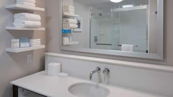 Un baño con lavamanos blanco, dos llaves, un espejo y estantes con toallas dobladas y artículos de aseo.