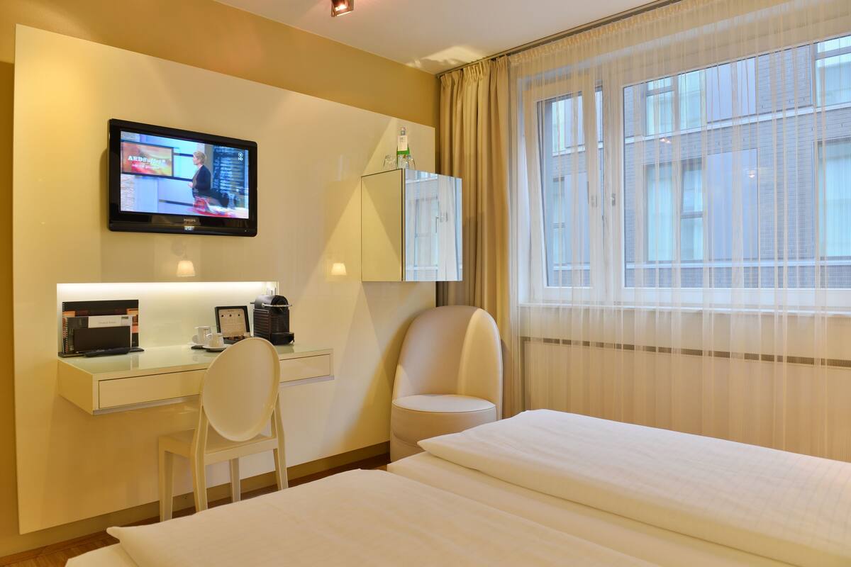 Une chambre d’hôtel avec un lit, un bureau équipé d’un ordinateur, une chaise, une télévision et une fenêtre avec des rideaux.
