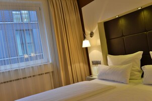 Una habitación de hotel con una cama grande, cabecero, mesita de noche, lámpara y ventana con cortinas.