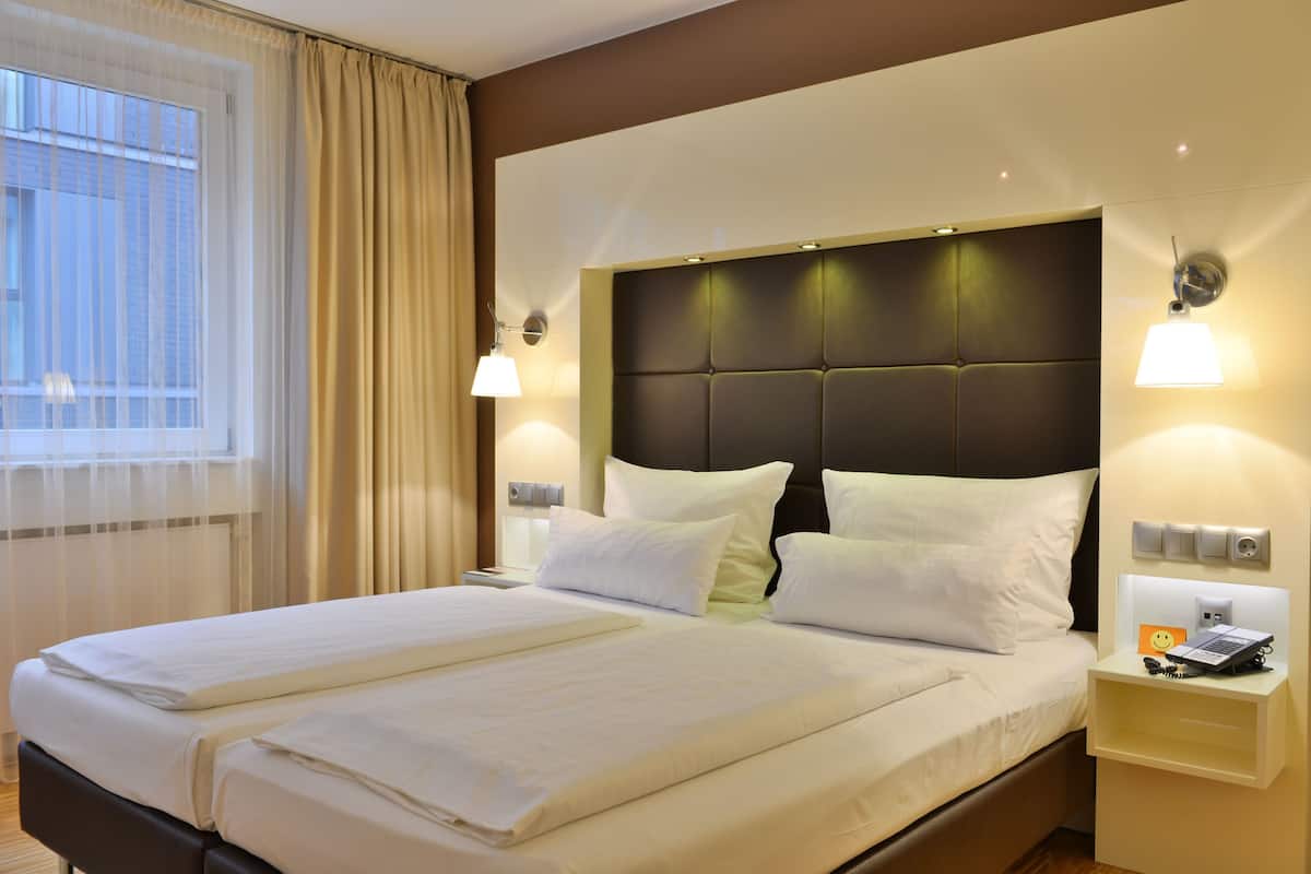 Une chambre d’hôtel avec un grand lit, deux tables de chevet, une tête de lit dotée d’un éclairage intégré et une fenêtre avec des rideaux.