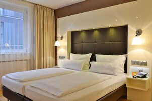 Habitación de hotel con una cama grande, dos mesitas de noche, una lámpara y una ventana con cortinas.