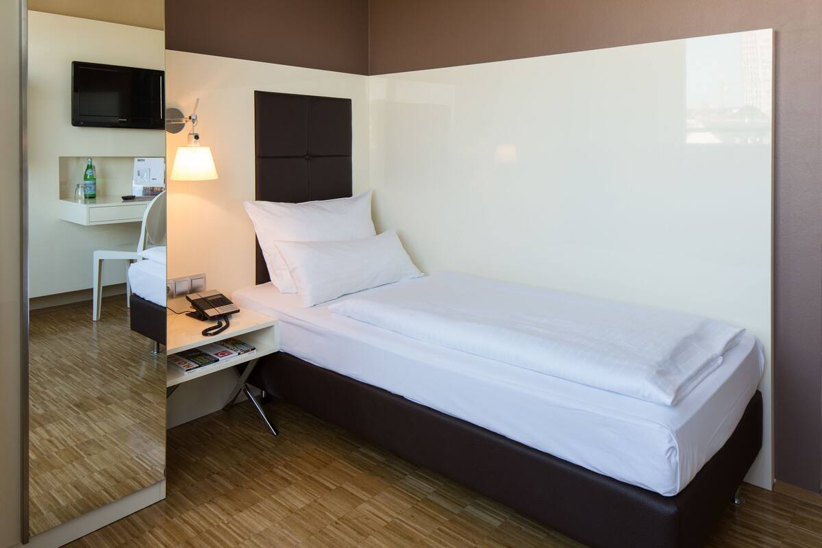Une chambre d’hôtel équip ée d’un lit, d’une table de chevet, d’une lampe, d’un bureau, d’une chaise et d’un téléviseur à écran plat fixé au mur.