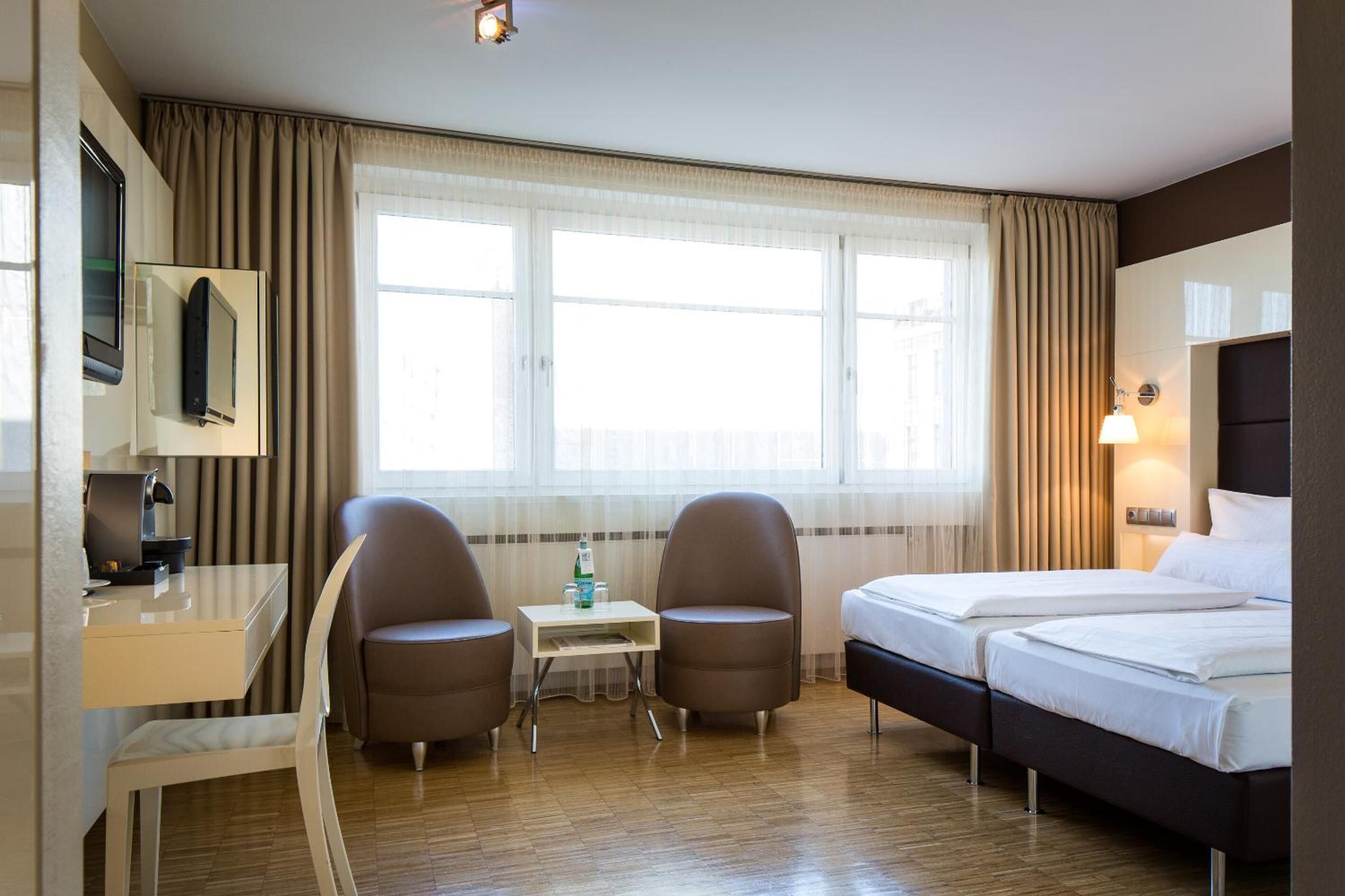 Una habitación de hotel con cama, escritorio, dos sillas, televisor y ventana con cortinas.