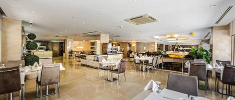 A spacious hotel dining area with multiple tables and chairs, a central bar, and a large chandelier.