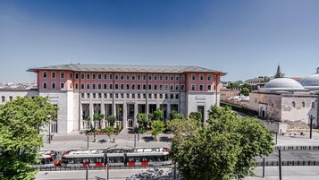 A multi-story building with a pink facade and large windows, surrounded by trees and a tram line.