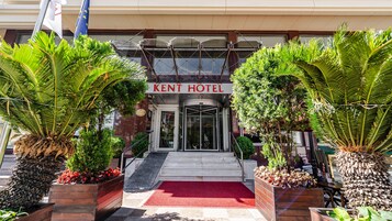 La entrada del Hotel Kent, con puerta giratoria, alfombra roja y plantas tropicales.