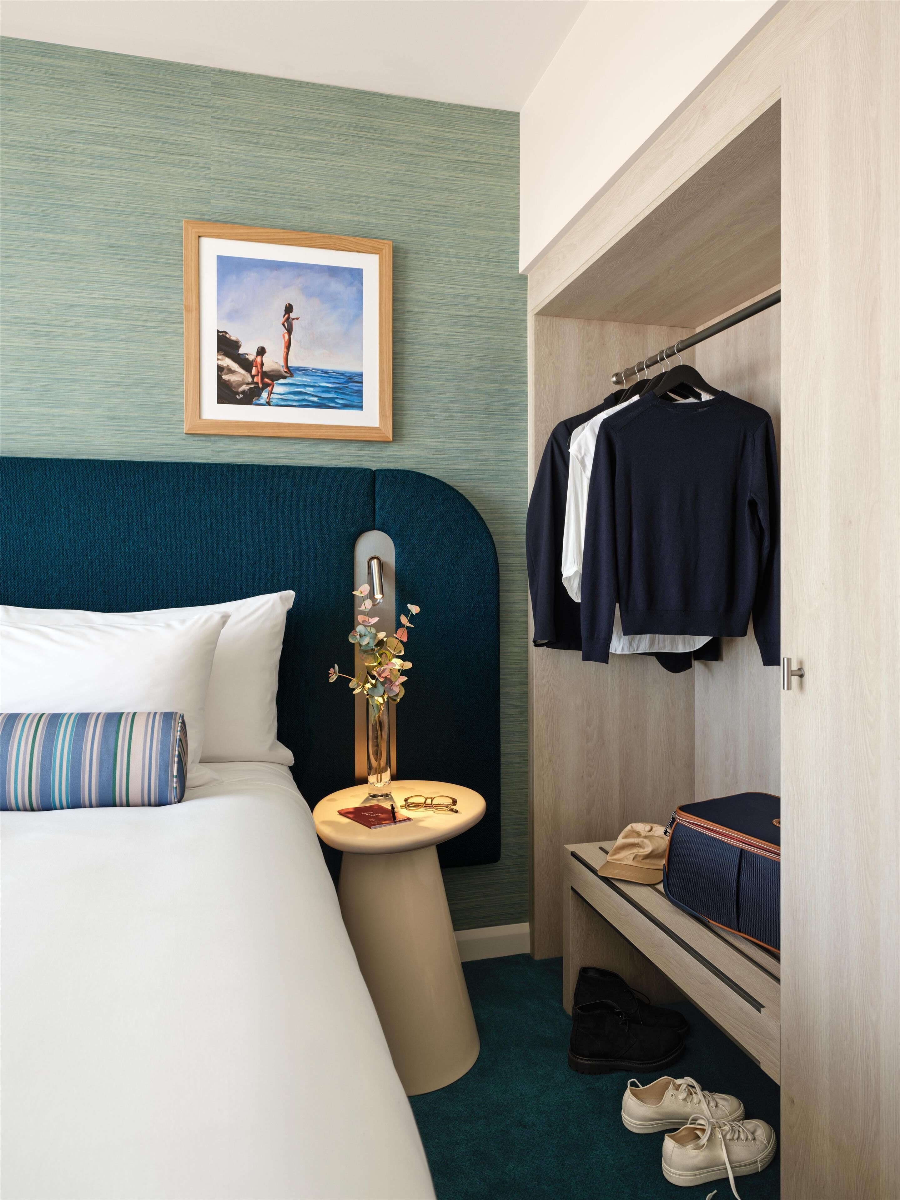Habitación de hotel con cama, mesita de noche con un florero y vasos, armario con ropa y un cuadro en la pared.