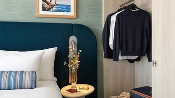 Habitación de hotel con cama, mesita de noche con un florero y vasos, armario con ropa y un cuadro en la pared.