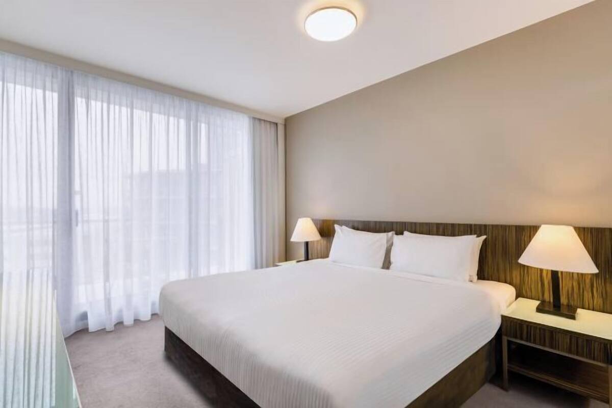 A hotel room with a large bed, two bedside tables with lamps, a headboard, and a large window with sheer curtains.