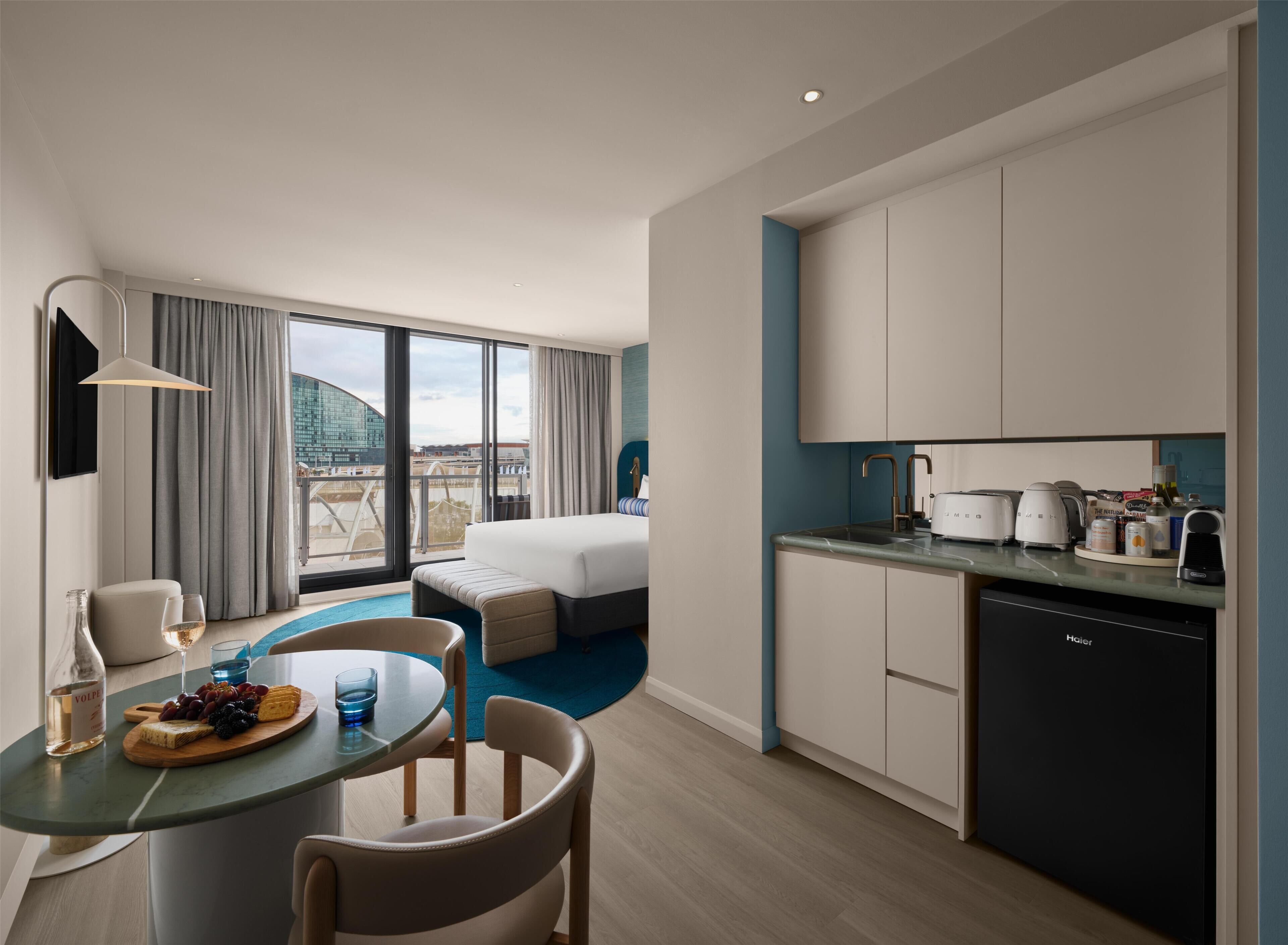 A modern hotel room with a bed, a dining area with a table set for two, a kitchen with a dishwasher, and a large window with a city view.