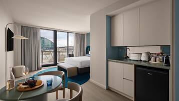 A modern hotel room with a bed, a dining area with a table set for two, a kitchen with a dishwasher, and a large window with a city view.