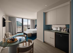 Habitación de hotel moderna con cama, zona de comedor con mesa para dos, cocina equipada con lavavajillas y un amplio ventanal con vistas a la ciudad.