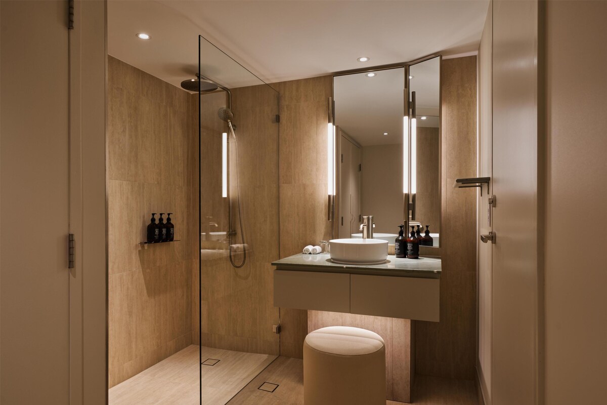 Un baño moderno con ducha de vidrio, tocador con espejo y un taburete redondo.