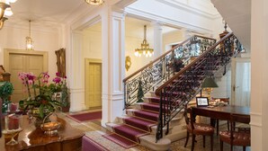 A grand staircase with ornate metal railings, a wooden table with chairs, and a vase of pink orchids.
