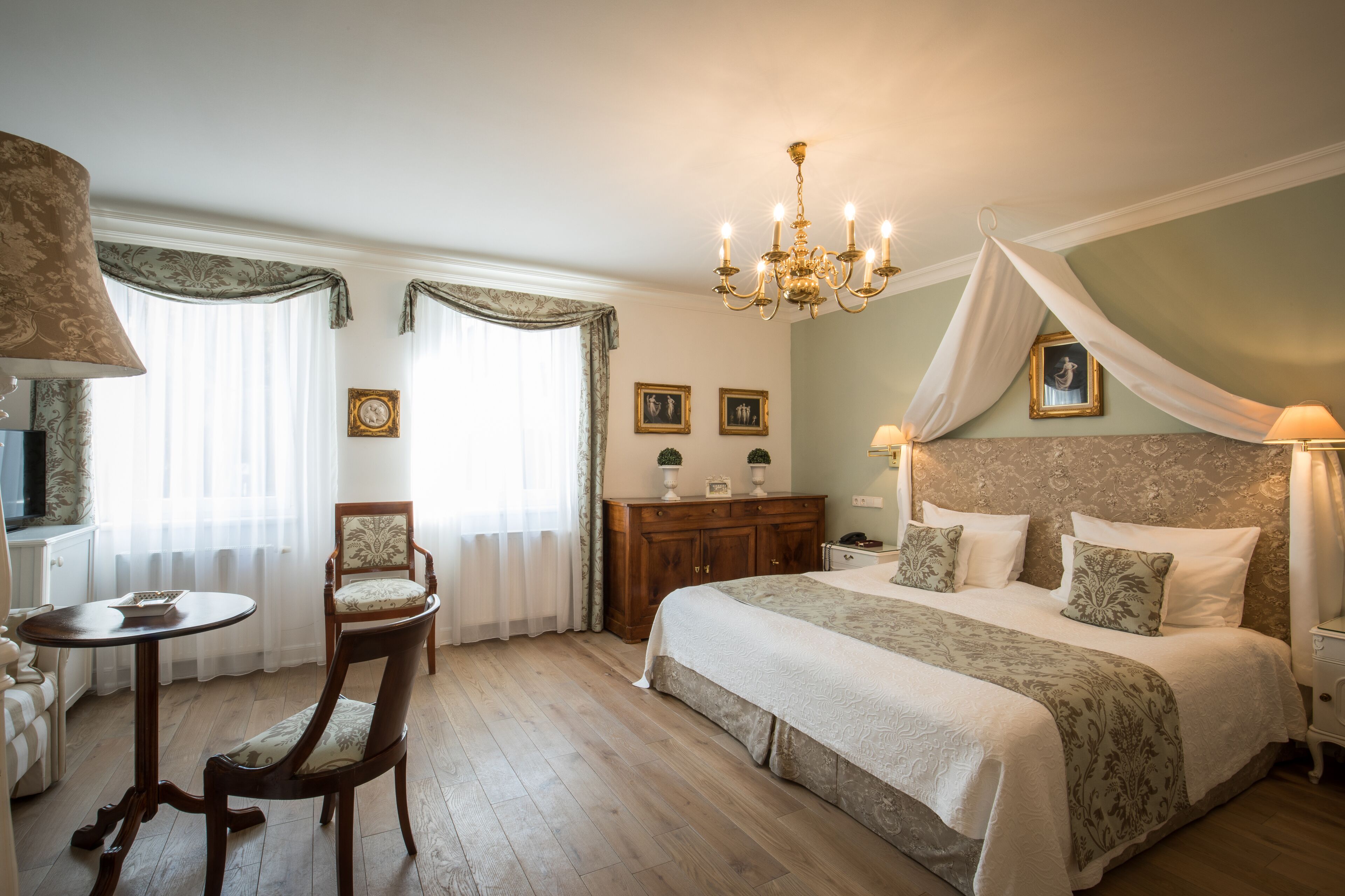 Quarto com uma cama grande, uma cadeira de madeira, uma mesinha e um candelabro.