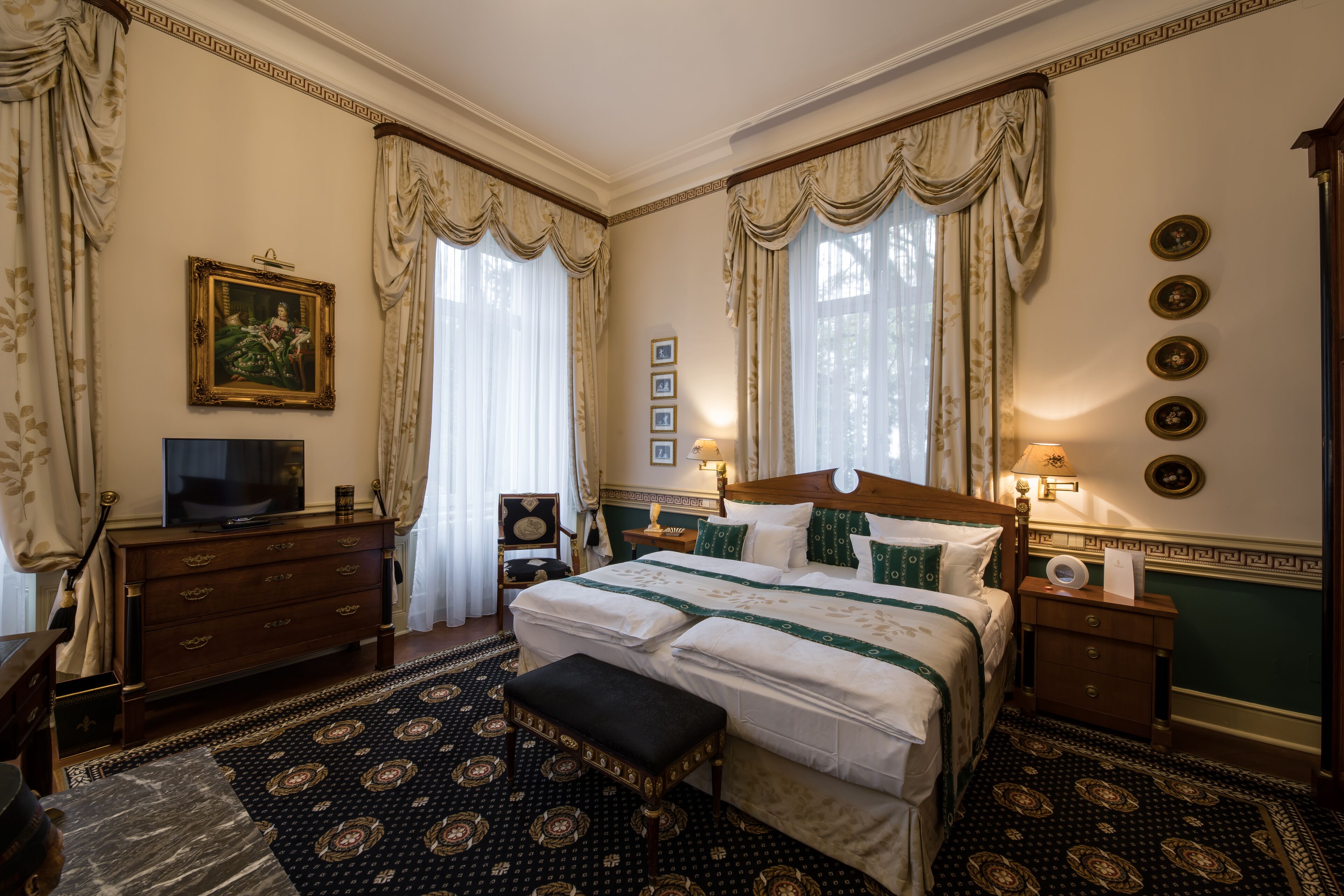 A classic, elegantly furnished bedroom with a large bed, a dresser, a television, and a bedside table with a lamp.