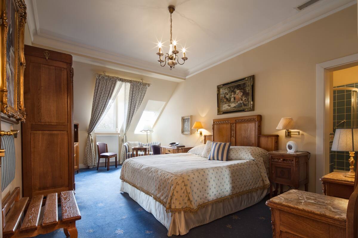 A bedroom with a large bed, a wooden bench, a desk, a chair, and a chandelier.