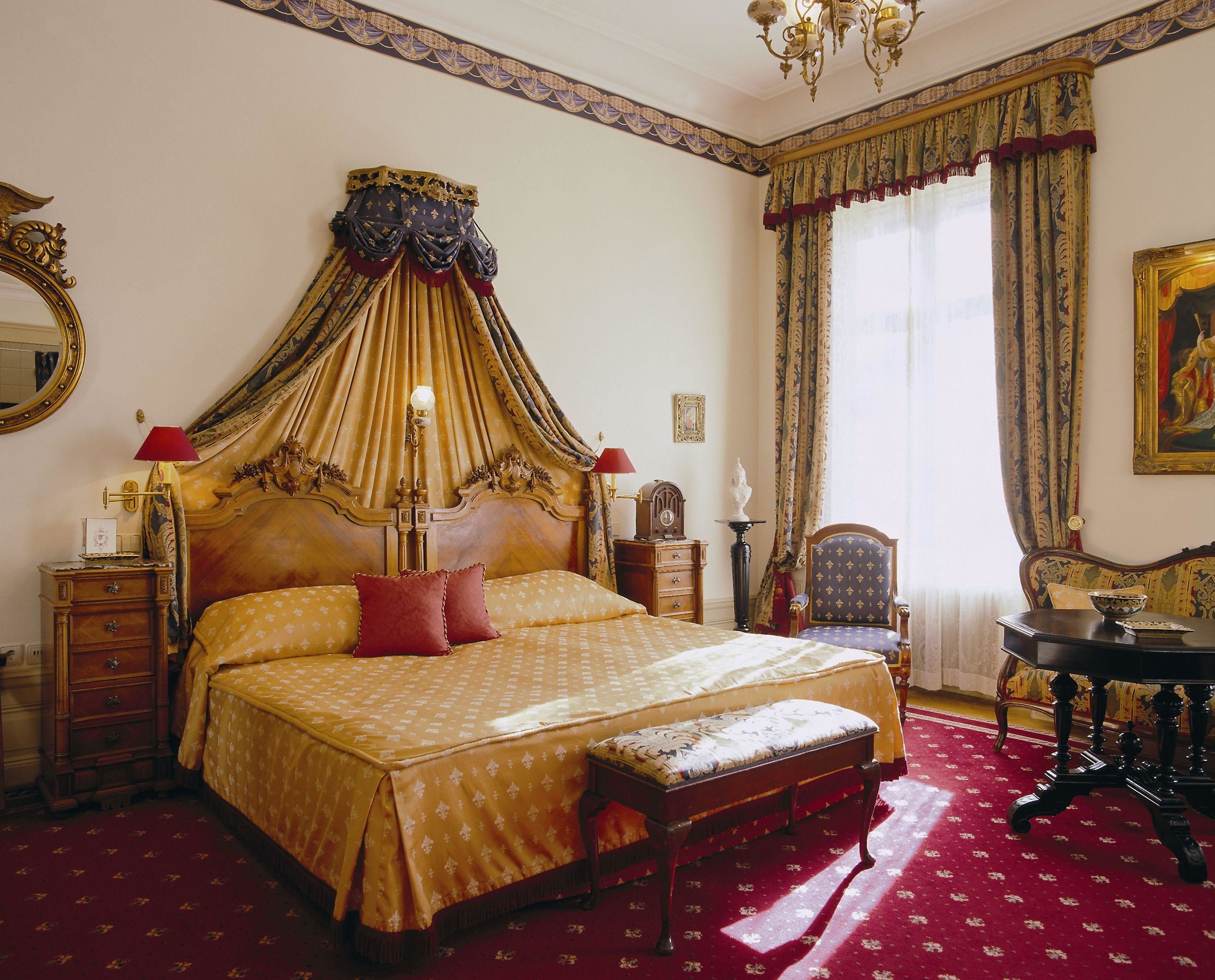 A large, ornate four-poster bed with a canopy, a wooden nightstand, a chair, and a table with a vase.