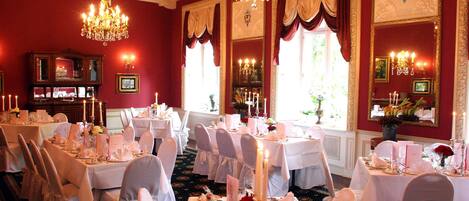 Un comedor formal con mesas dispuestas para un banquete, candelabros y amplias ventanas con cortinas.