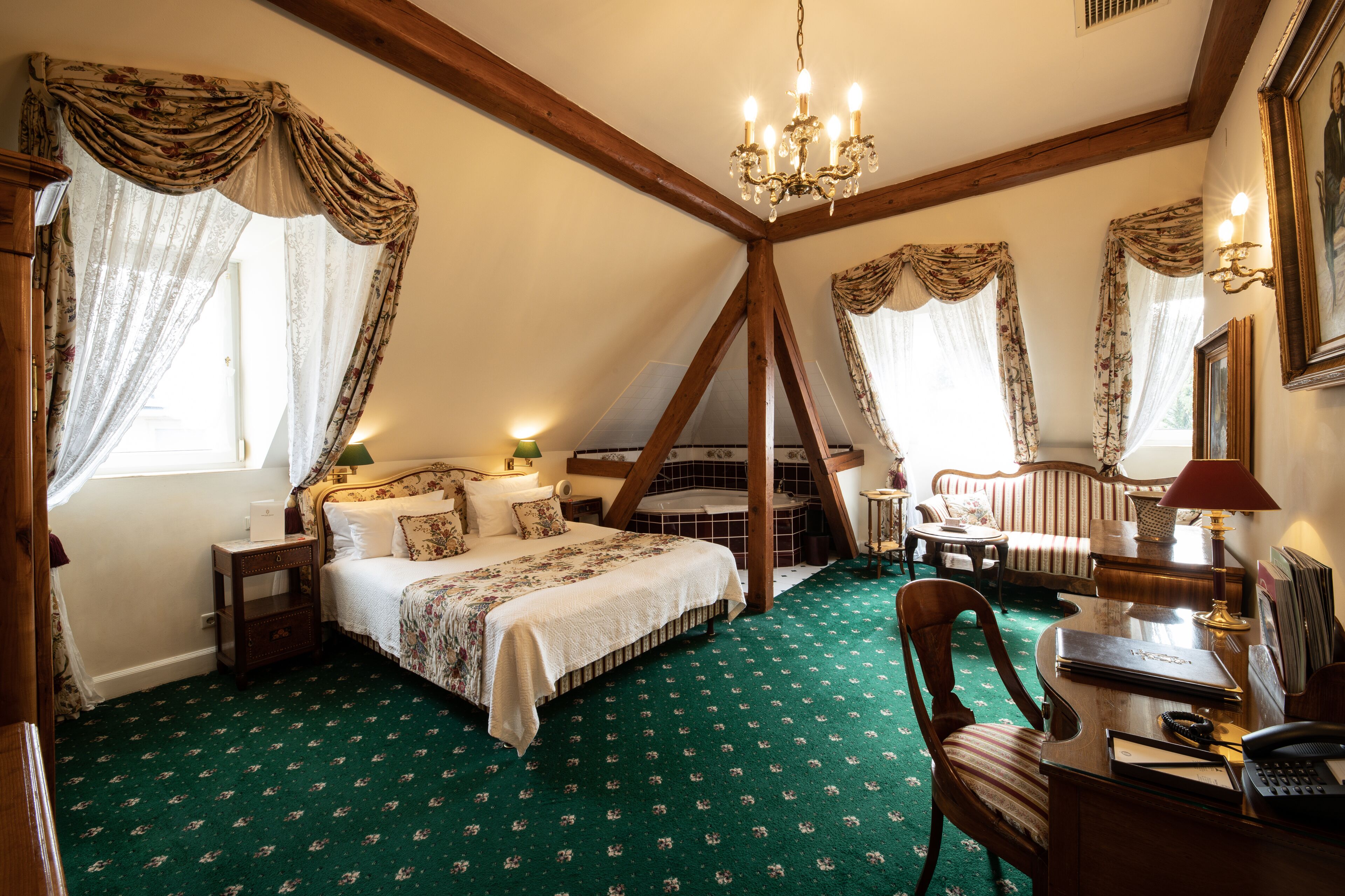 A bedroom with a large bed, a desk with a chair, a small table, and a chair. There is a chandelier hanging from the ceiling.
