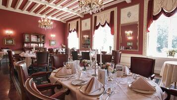 Uma sala de jantar formal com mesas postas, lustres e quadros nas paredes.