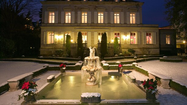 A grand building with a central fountain, surrounded by snow-covered garden beds and trimmed hedges.